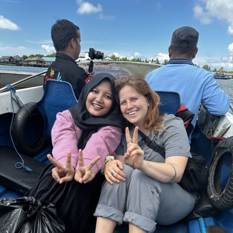 PhD student sits on a boat with an Indonesian woman, holding up peace signs