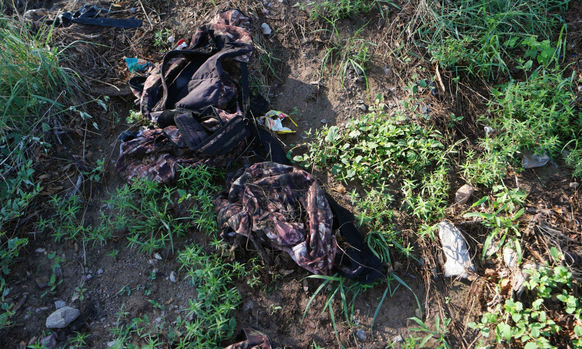 Camouflage backpacks and discarded clothing scattered on the ground among dirt and plants.