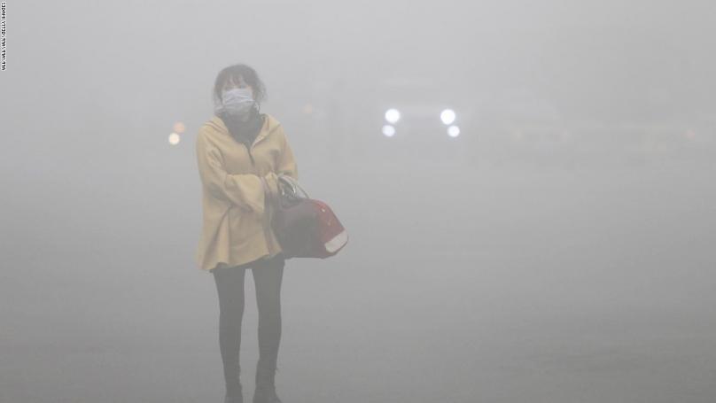 Amidst dense smog in a Chinese urban setting a woman stands alone wearing a mask