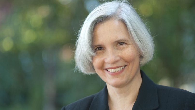 Headshot of Shelley Rigger. She is an older woman with silver gray hair cut in a bob style wearing a black top. She faces the viewer with a smile. Background is out of focus green foliage.