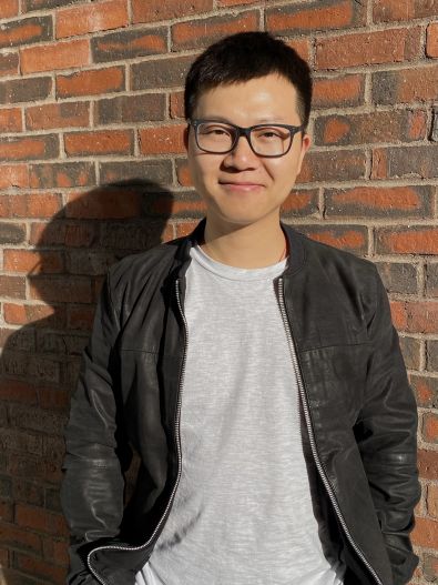 Wanheng Hu stands in front of a brick wall wearing glasses, dark leather jacket and a gray t-shirt. His hair is short cropped and black. He is Chinese.
