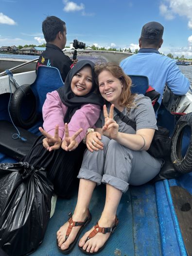 PhD student sits on a boat with an Indonesian woman, holding up peace signs