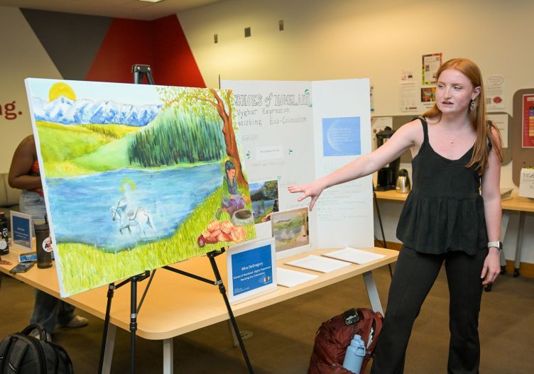 Mira DeGregory gestures toward her painting of a person seated in grass by a lake. 
