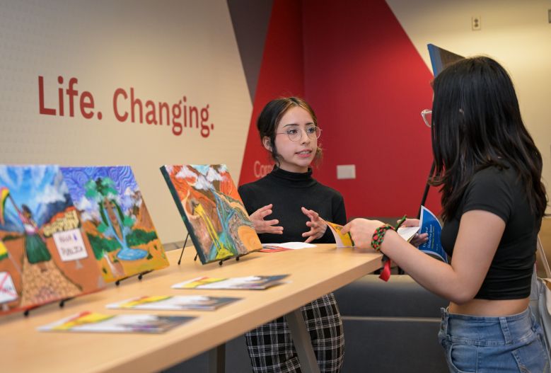 A Global Scholar talks with their hands to another student, standing alongside a final art project.