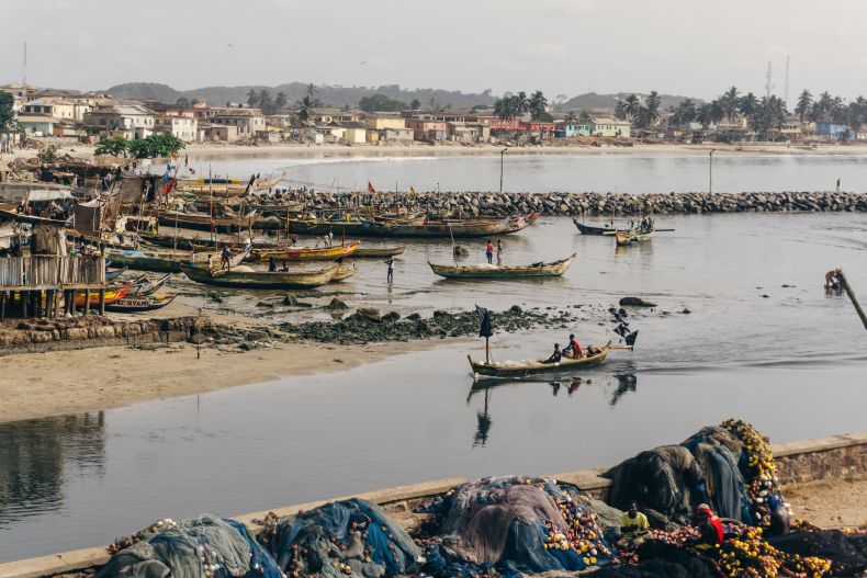 Long, slim boats move down a shallow river, surrounded by coastal housing and working fishers