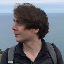 Thomas wears longish brown hair backpack and dark shirt facing in profile with a blue sea background