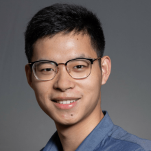 Tianli wears glasses and smiles at camera wearing a blue shirt against a gray backdrop