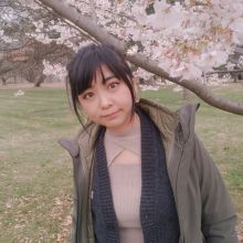 Yumeng leans under a bough of flowering cherry trees with long pulled back hair a gray jacket dark sweater and pink blouse