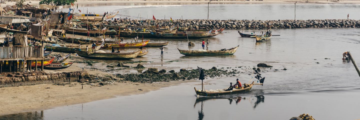 Long, slim boats move down a shallow river, surrounded by coastal housing and working fishers