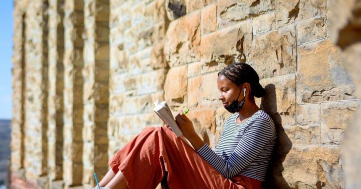 A Cornell student leans against a wall on campus and writes in a journal.