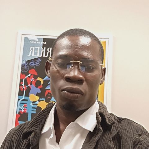 Man in collared shirt and jack stands in front of white wall and poster. 