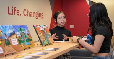 A Global Scholar talks with their hands to another student, standing alongside a final art project.